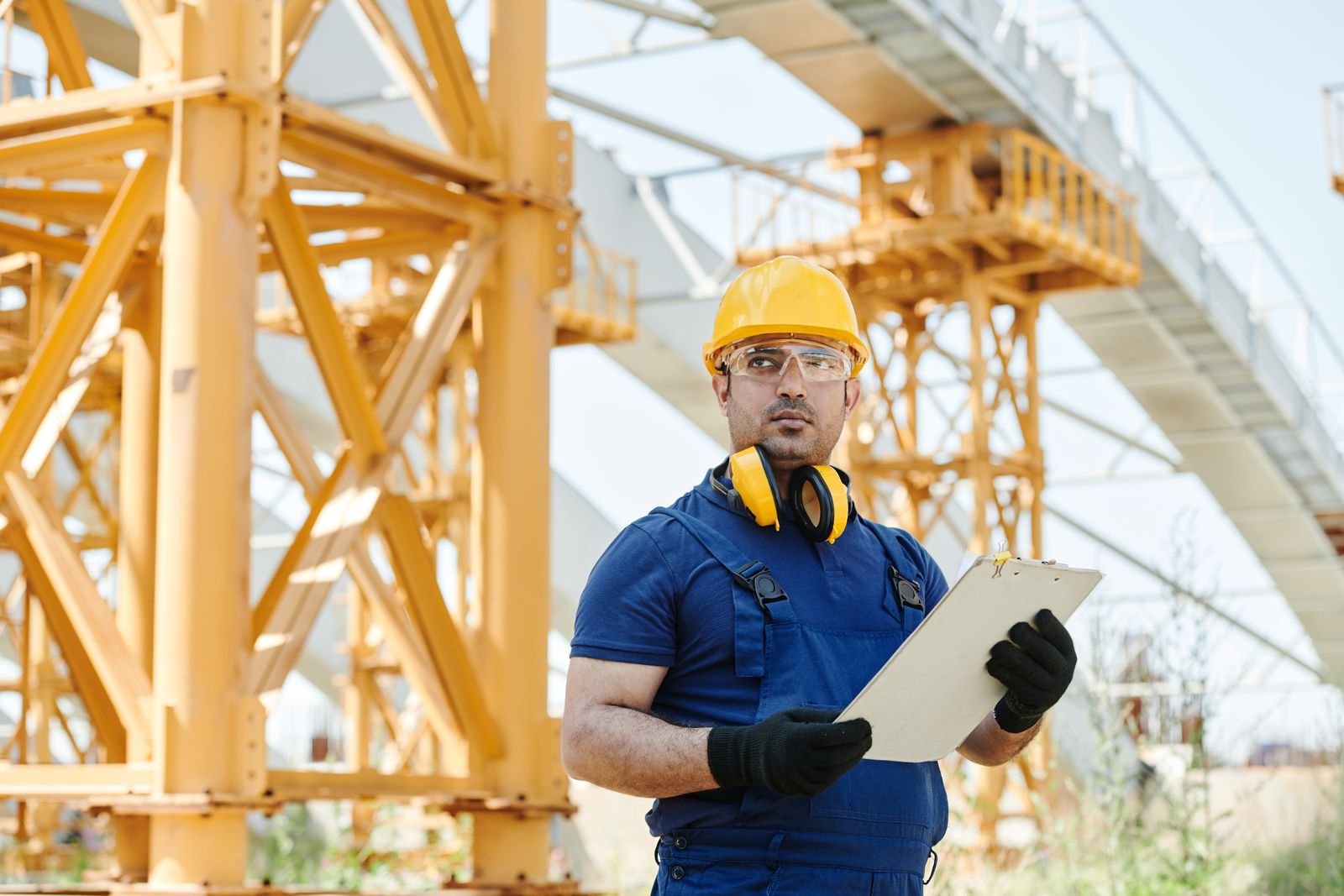 Worker in safety gear inspecting a construction site with a clipboard.