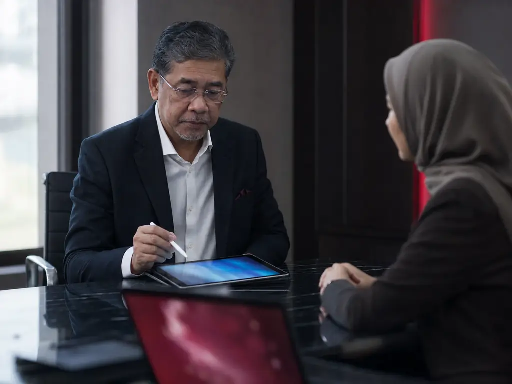A senior manager pausing with a stylus above a tablet, deciding whether to approve, while a colleague waits opposite.